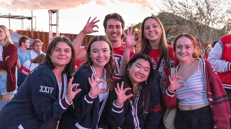 A group of University of Arizona students stand together outside in front of a mountain, and each holds up the Wildcat hand symbol.