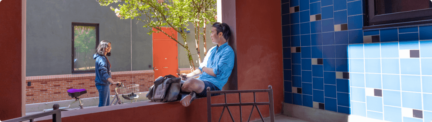 Person with grey hair sits on a wall chatting with younger person walking a bike